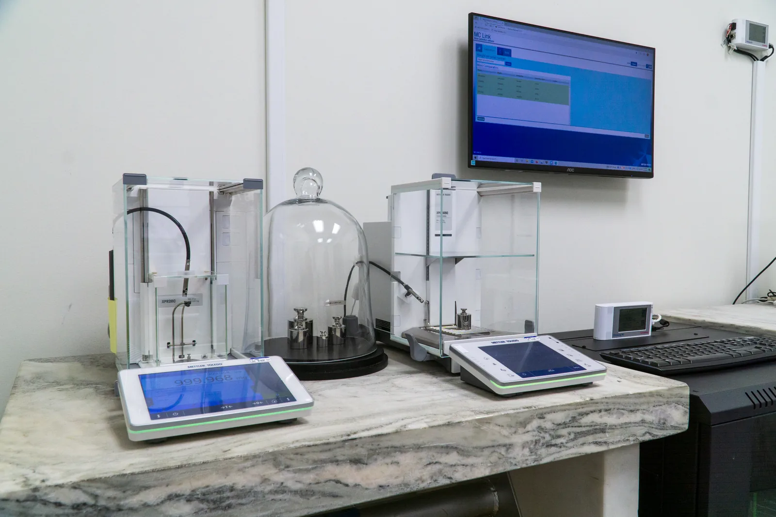 Mettler Toledo mass comparators on a stone isolation bench at American Gage, with reference weights under a bell jar and MCLink comparator software running alongside